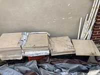 Stacks of beige/light tan 18x18 inch square tiles, stacked outdoors on plastic crates and other materials against a wall, showing some dirt and chips.