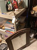 Wooden framed mirror with floral carving at top detail, leaning against shelving unit with books and other items behind