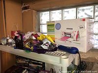 Wide view showing boxed Cricut machine, bottles, crafting materials, and flags on a white plastic shelf against a concrete window wall