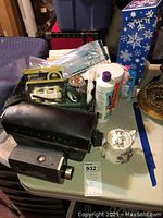 Bell & Howell Super 8 camera with black carrying case, assorted crafting items including spools of thread and a small ceramic miniature teapot, white bottle with purple lid, and metal circular object, alongside a tile in blue snowflake box on a table.