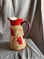 Photo of hand-painted ceramic pitcher with large red poppy flowers on beige background and red rim and handle.
