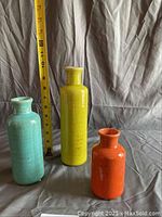 Photo showing three ceramic vases of varying heights and colors (turquoise, yellow, orange) with a measuring tape for size reference