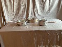 Photo of three stainless steel sauce pans with glass lids displayed on a cloth-covered surface, showing different sizes and long handles.