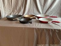 Set of five frying pans displayed on a beige surface and beige background, showing assortment of materials and colors including non-stick and cast iron.