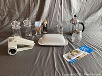 Photo of assorted kitchen items displayed on gray fabric background including butter dish, French press, grinders, shakers, toothpick holders, and handheld tool.