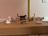 All six cat figurines displayed on a cardboard surface with a ruler behind them showing their relative height and size.