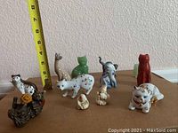 Nine assorted cat figurines on a flat surface, including a measuring tape showing height of approximately 7 inches for one tall figurine.