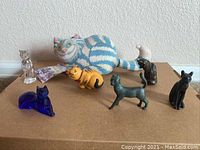 Eight assorted cat figurines, varying materials including plush, crystal, metal, ceramic, and glass. Includes large plush cat, crystal cat, blue glass cat, bronze metal cat, black ceramic cat, orange tiger-striped ceramic cat, mottled ceramic cat, and a purple decorative glass piece.