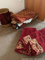 Photo of stacked red and gold decorative hand towels, bath mat, trash can, and soap dispenser together on carpeted floor.