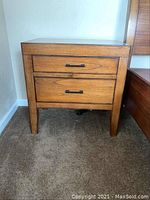 Front view of one solid wood nightstand showing two drawers and tapered legs on carpeted floor by bed frame