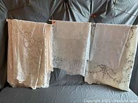 Various lace cloth pieces hanging over a bar, showing detail of texture and patterns, including floral and ornamental embroidered designs in cream and white colors.