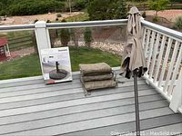 Patio umbrella with metal pole, folded beige umbrella canopy, box containing patio umbrella base stand, and four beige patio cushions stacked and tied together placed on deck against railing.