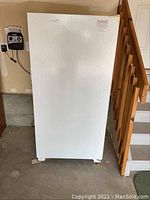 Closed front view of white upright garage refrigerator next to wooden staircase, placed on concrete garage floor.