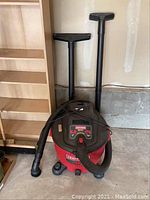 Photo showing red and black Craftsman 6.0 HP shop vacuum with hose attached and two plastic nozzle accessories standing against garage wall.