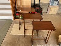 Photo shows three wooden TV trays arranged on a garage floor near stairs and storage items. Trays are rectangular with folding legs and medium brown wood finish, showing wear.