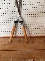 Manual hedge clippers with wooden handles displayed against a pegboard wall and resting on a wooden surface, showing wear on handles and metal blades.