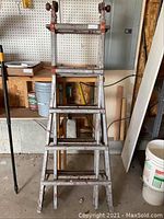 Metal extension ladder shown upright leaning against a workbench in a garage setting, with paint splatters and worn spots visible on the rungs and side rails.