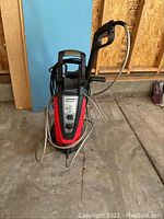 Full view of the Husky pressure washer showing the unit with attached hose and spray gun, placed on a concrete floor with a wooden wall in the background.