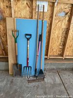 Full view of the five garden tools leaning against a wall in a garage: shovel, pitchfork, two rakes, and a hoe