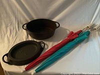All items displayed on white cloth: cast iron pan with lid and two umbrellas (red and turquoise).