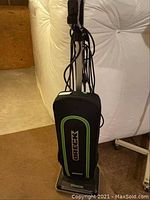 Front view of green and black Oreck upright vacuum cleaner with fabric bag and electrical cord wrapped around upper hooks.