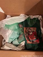 Photo of an unboxed Santa Claus figurine wrapped in green bubble wrap and tissue paper inside a cardboard box with a Santa image on the wrapping material.