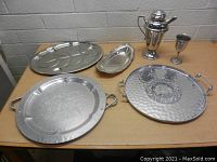 Photo of six metal serveware pieces on a wooden table against a brick wall background including assorted platters, a pitcher, and a goblet.