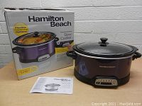 Hamilton Beach programmable slow cooker in purple with glass lid, original box featuring cooking image with food inside, instruction manual visible, control panel with buttons and digital display on base.