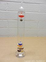 Full view of the glass Galileo thermometer showing the tall cylindrical shape, clear liquid, and multiple colored bulbs floating inside.