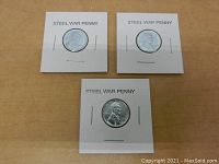Three 1943 steel war pennies each mounted in cardboard holders labeled STEEL WAR PENNY placed on a wooden surface.