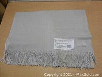 Folded taupe wool throw laid on flat surface showing fringed edge and two tags attached.
