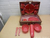 Vintage picnic basket open showing the red plastic dishware including plates, mugs, bowls, pitcher, and wrapped plastic cutlery arranged inside and outside basket.