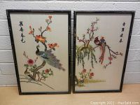 Pair of framed embroidered pictures under glass against white brick wall, showing bird and floral motifs with Chinese text.