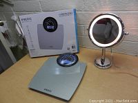 HoMedics digital bathroom scale with round digital display, box in background, and chrome lighted makeup mirror