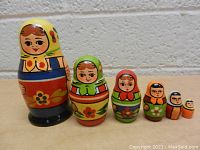 Six small wooden Russian nesting dolls lined up by size on a carpeted floor against a white textured wall.
