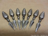 Seven vintage silver plated corn cob holders arranged in a row showing front view with corn cob shaped handles and pointed prongs.