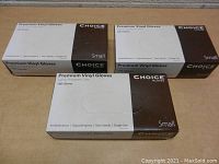 Three unopened boxes of Choice Premium Vinyl Gloves Size Small stacked on a wooden surface.