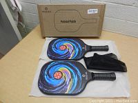 Two Ticci pickleball paddles with colorful swirl design, black grips, black protective covers, and the original box placed on a wooden table.