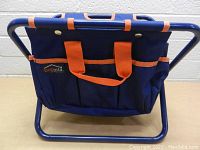Foldable chair with blue fabric tool bag and orange straps, metal tubular blue frame shown folded and upright on a table.
