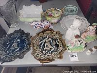 Table with varied vintage porcelain items including ornate plates, figurines, teacup, and decorative small dishes. Colors range from dark blue and gold to pastel floral and white porcelain.