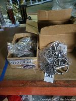 Photo showing two open cardboard boxes containing various metal belt buckles, some wrapped in plastic bags, displayed on a table with trophies in the background.