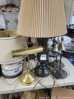 Photo showing large table lamp with beige pleated shade, brass desk lamp, ceramic lamp with blue and white design, and lamp with silver shades