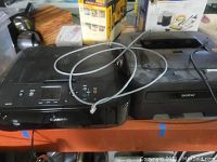 Photo showing front and top view of black Canon MG5720 printer next to a black Brother HL-2270DW printer on a table with gray cable across both devices.