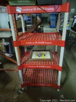 Oblique front view of the red and white Coca-Cola branded plastic shelving unit with four shelves and a framed decorative item on one shelf.