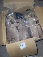 Open cardboard box filled with multiple bundles of tan suspenders bundled with rubber bands. Metal clips are clearly visible attached to the ends of the suspenders.