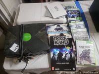 Photo showing original Xbox console with attached AV cables, Nintendo Wii console with sensor bar and manual, multiple Xbox 360 video games, and Rock Band guides.
