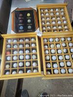Four display cases of golf balls arranged together, three wooden cases with multiple compartments each holding mostly white golf balls, one smaller framed case with six colored golf balls.