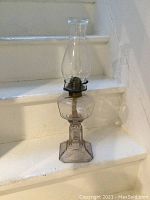 Full view of the 19-inch tall clear glass hurricane lamp on stairs, showing entire lamp with octagonal base, central reservoir, metal wick holder, and chimney.