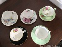 Five assorted fine bone china cups and saucers with varied floral designs and gold accents displayed on table