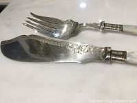 Close-up of a large serving knife and fork showing mother of pearl handles, sterling silver shanks with ridged collar accents, and silver plated blade and tines with scroll engraving.
